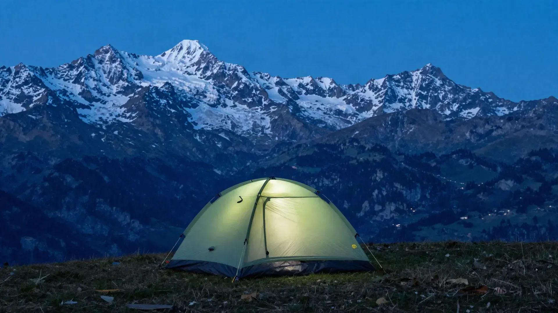 Ein beleuchtetes Zelt auf einer Alpwiese in der Dämmerung, dahinter verschneite Gipfel.